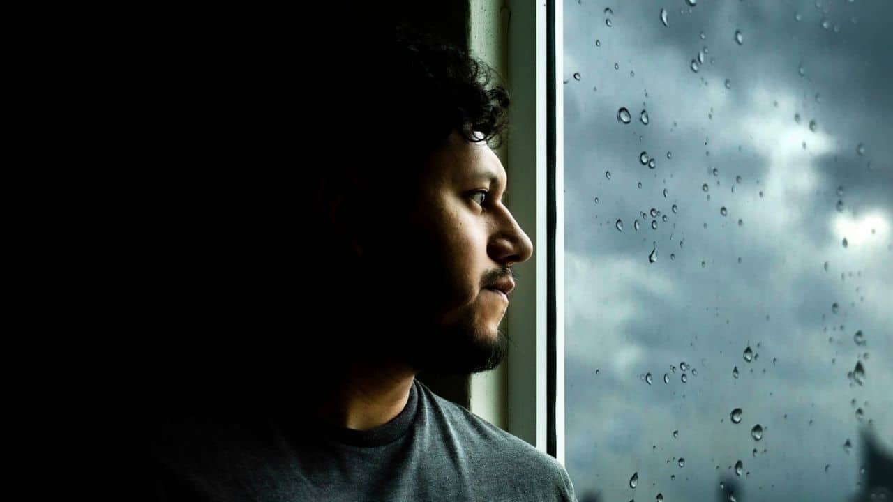 Man looking out a window covered in raindrops on a dark, cloudy day.
