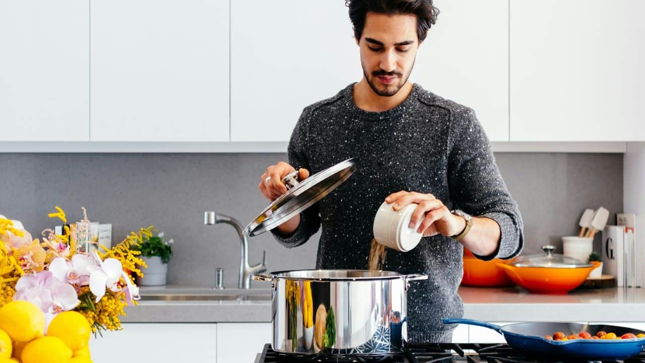 Man in a grey sweater cooking in a bright kitchen, adding ingredients to a pot.