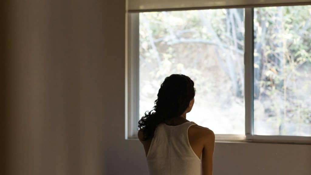 A person with long dark hair sits by a window, looking outside into the sunlight.