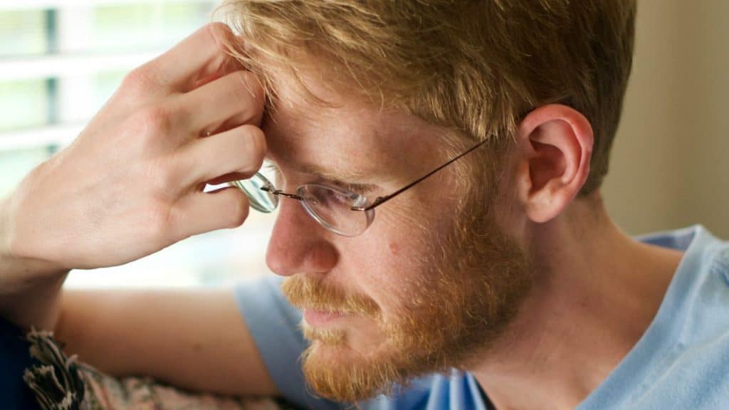 Close-up of a concerned man with a red beard and glasses, resting his hand on his forehead.
