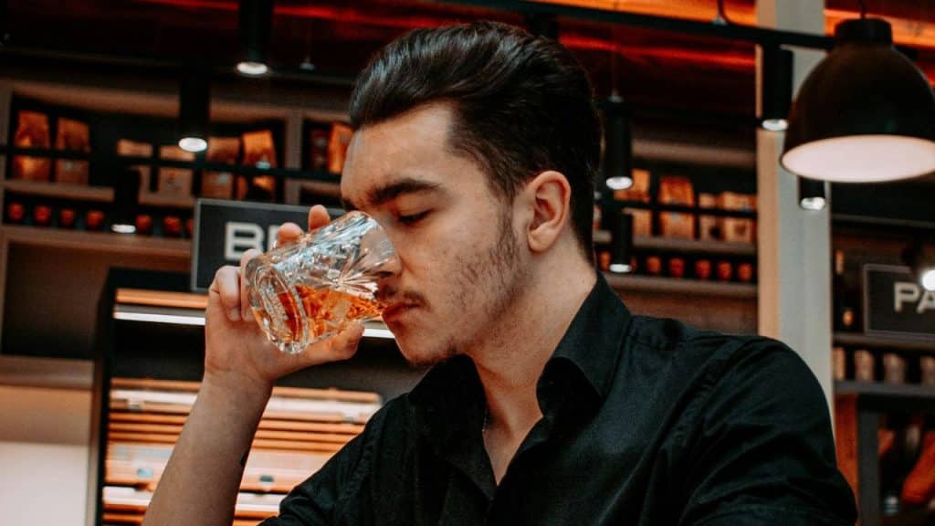 Young man in a black shirt taking a drink from a glass in a dimly lit bar or cafe.