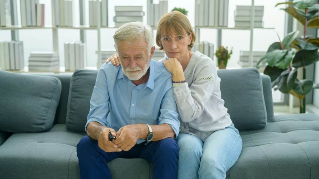 Elderly couple sitting sadly on a couch; woman's arm around man; bookshelf background.