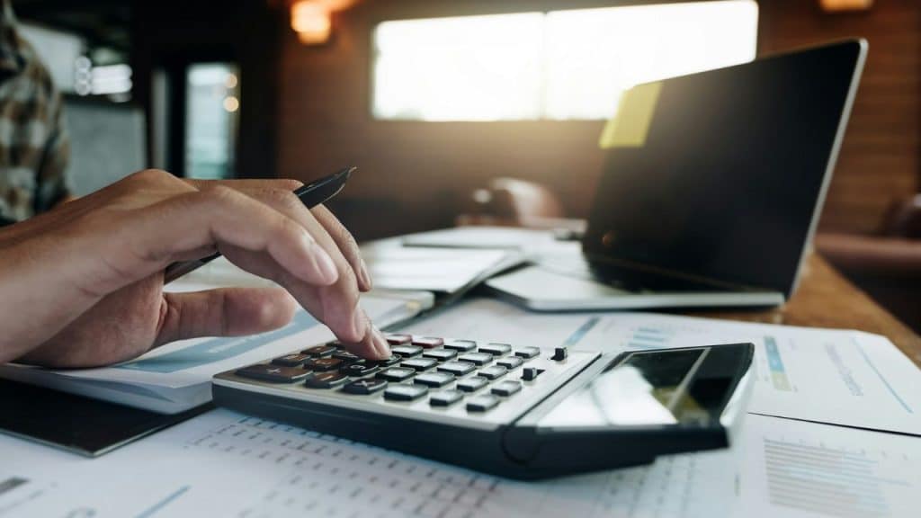 Close-up of a hand using a calculator on a desk with papers and a laptop.
