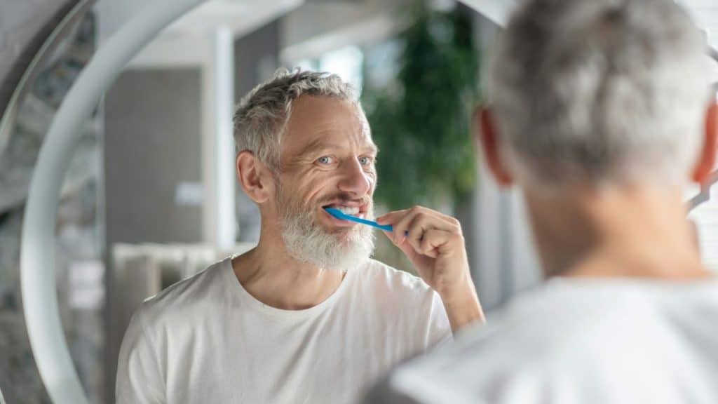 Middle-aged man with a gray beard brushing his teeth, looking in a mirror.