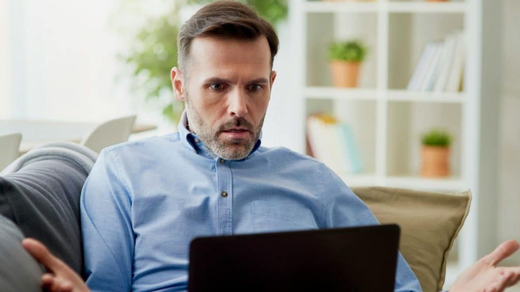 Man in a blue shirt sitting on a couch, looking stressed while using a laptop.