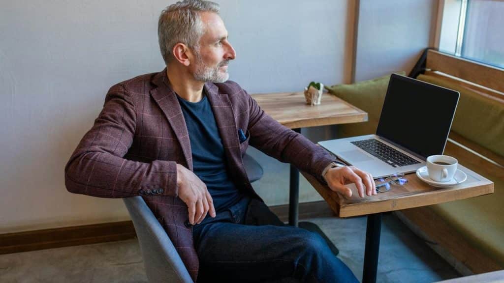 A man with gray hair sitting at a cafe table with a laptop and coffee, looking out the window.