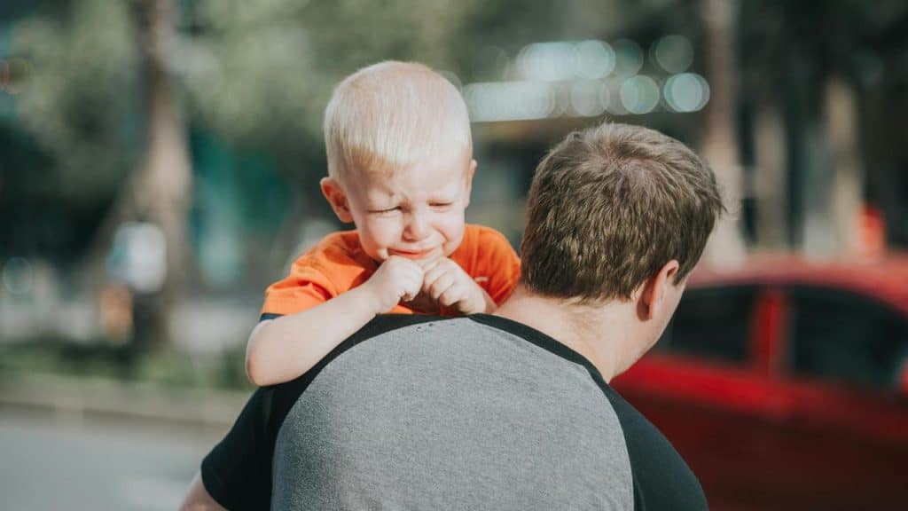 A man carrying a crying child on his shoulder outdoors.