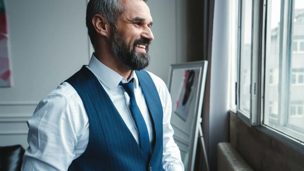 Smiling bearded man in a white shirt, blue vest, and tie looking out a window.