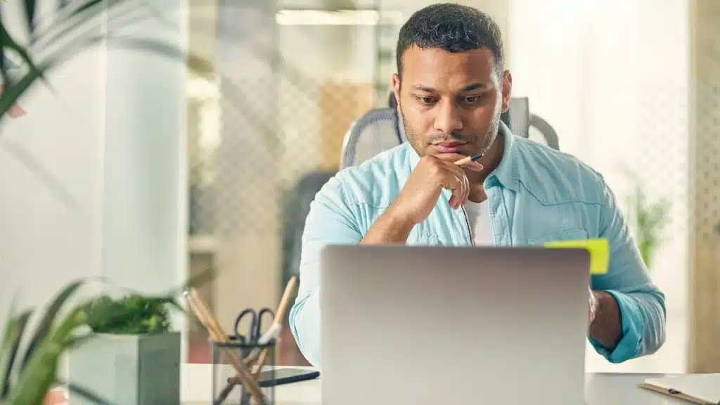 Man in a light blue shirt looking intently at a laptop, resting his chin on his hand.