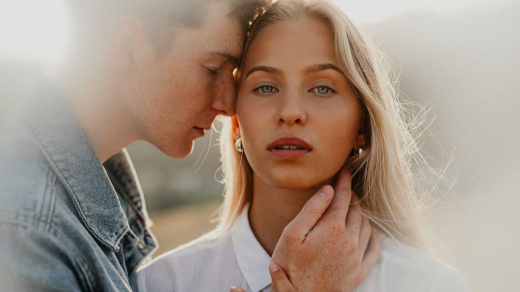 Young man with freckles leaning his head on a blonde woman's forehead, holding her face.