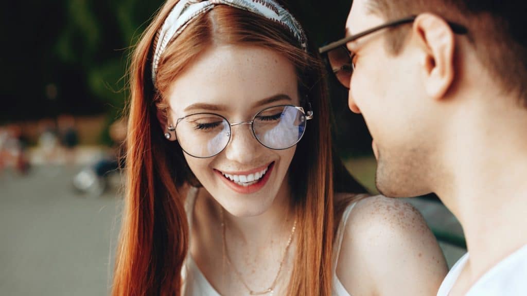 Close-up of a smiling redhead woman in glasses talking to a man.