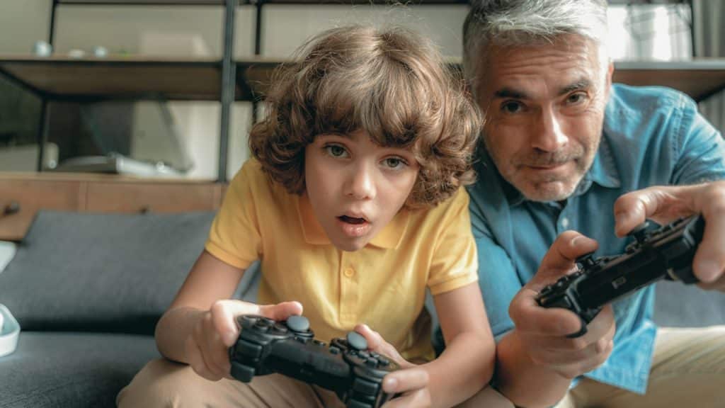 A little boy sitting near his father and playing a video game with him.