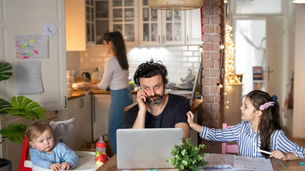 A man sitting at a table is working on a laptop and looking after his children simultaneously while his wife is working in the kitchen.