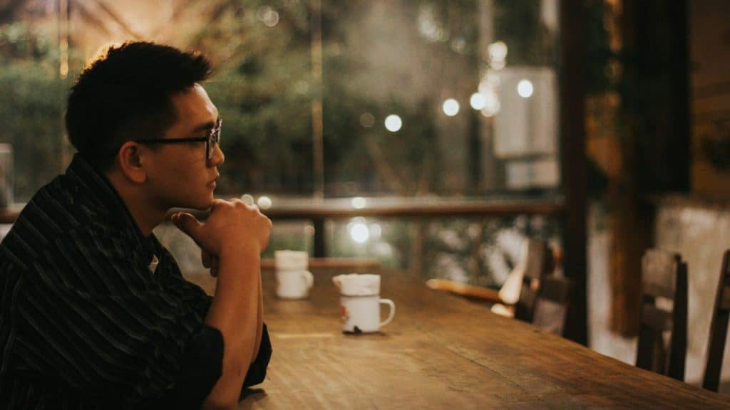 A man with glasses sits at a wooden table in a dimly lit space, chin resting on his hands.