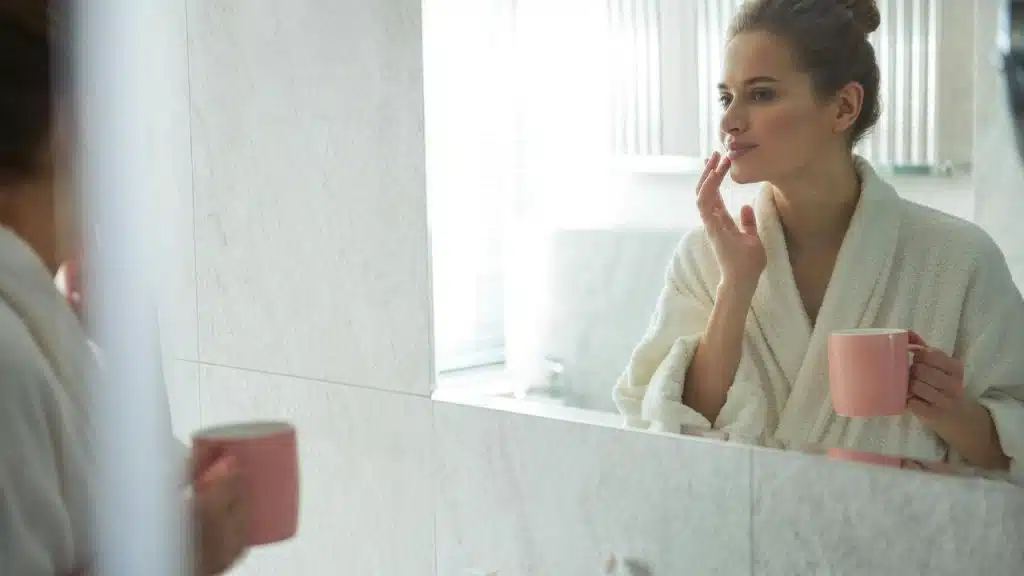 A woman in a bathrobe holds a pink mug while looking in the mirror and touching her lips.
