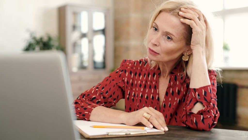 A blonde woman in a red patterned shirt looks intently at a laptop screen, hand to her head.