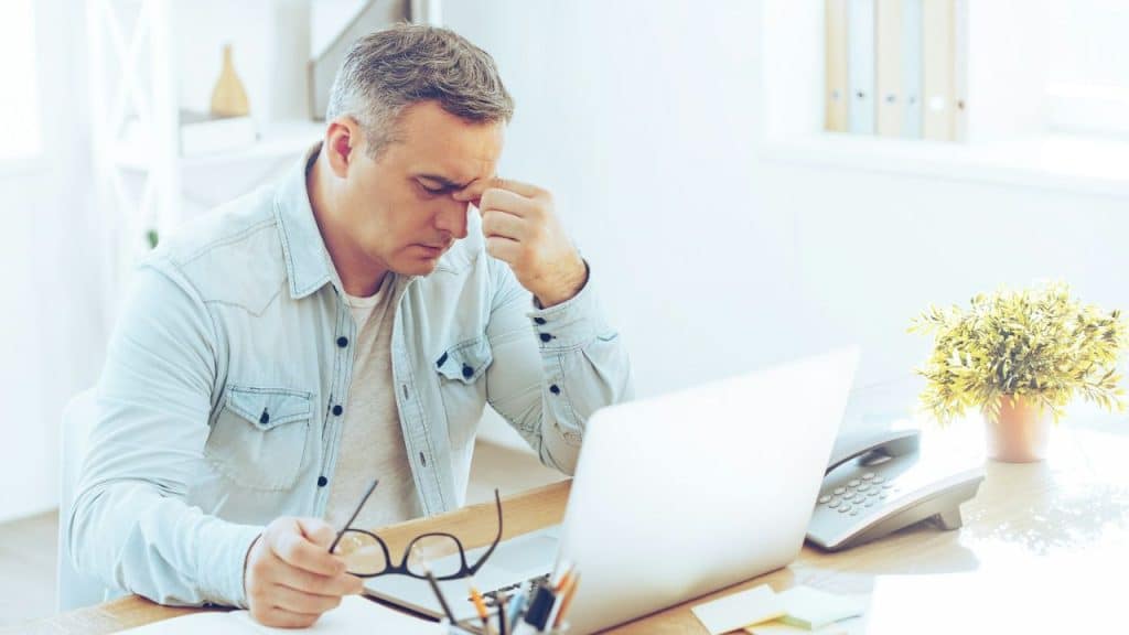 A man in a light blue shirt, holding glasses, sits at a desk rubbing his forehead.