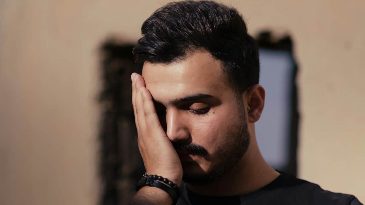 A man with dark hair, a beard, and a mustache covers his face with his right hand.