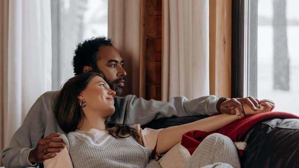 A couple relaxing on a couch while holding hands and looking out the window.