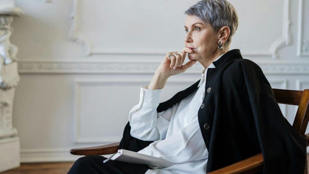 A thoughtful woman with short gray hair sits in a chair holding a notebook.\