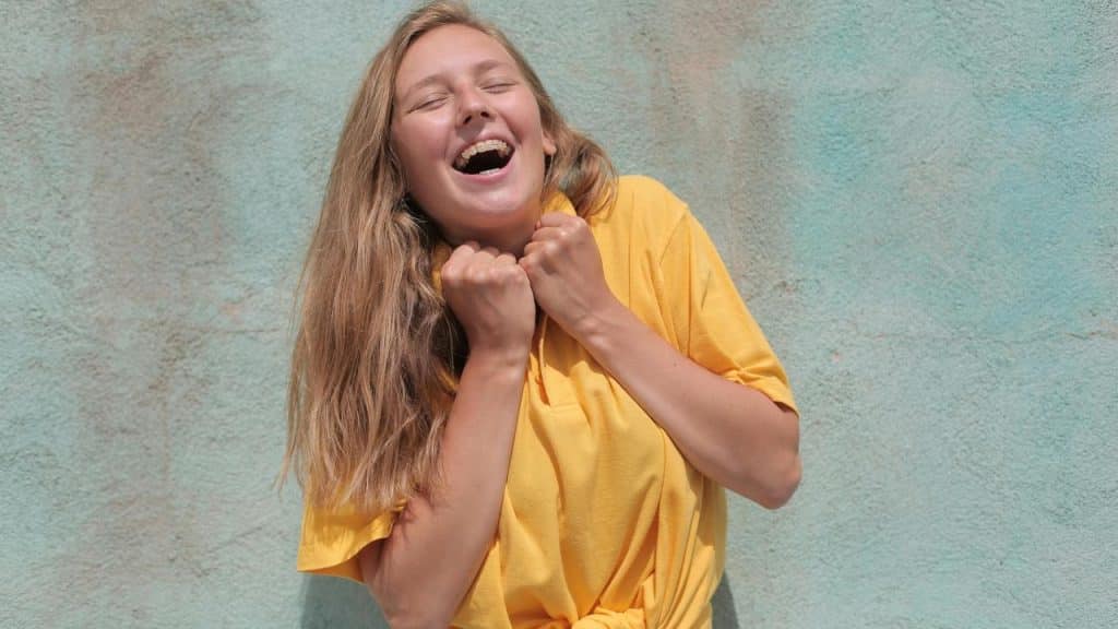 A joyful woman in a yellow shirt laughing with her hands clasped near her face.