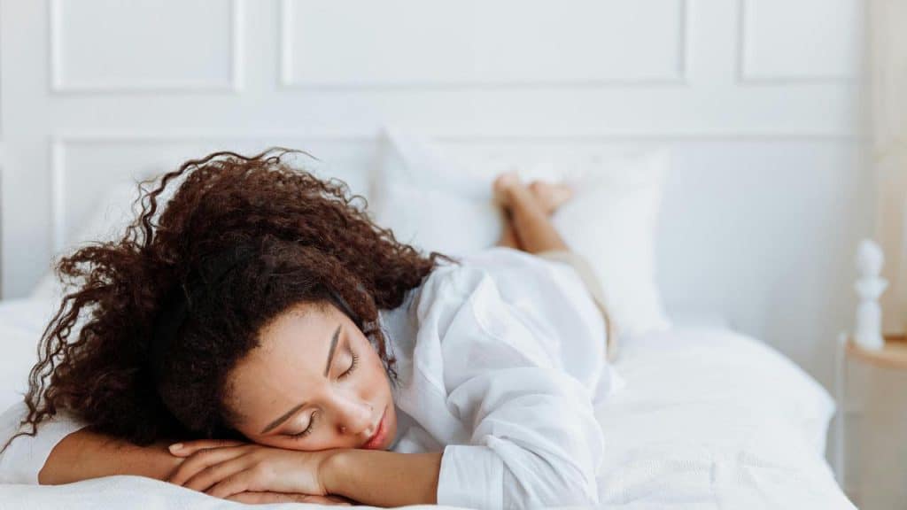 A woman sleeping on her stomach on a white bed in a bright bedroom.