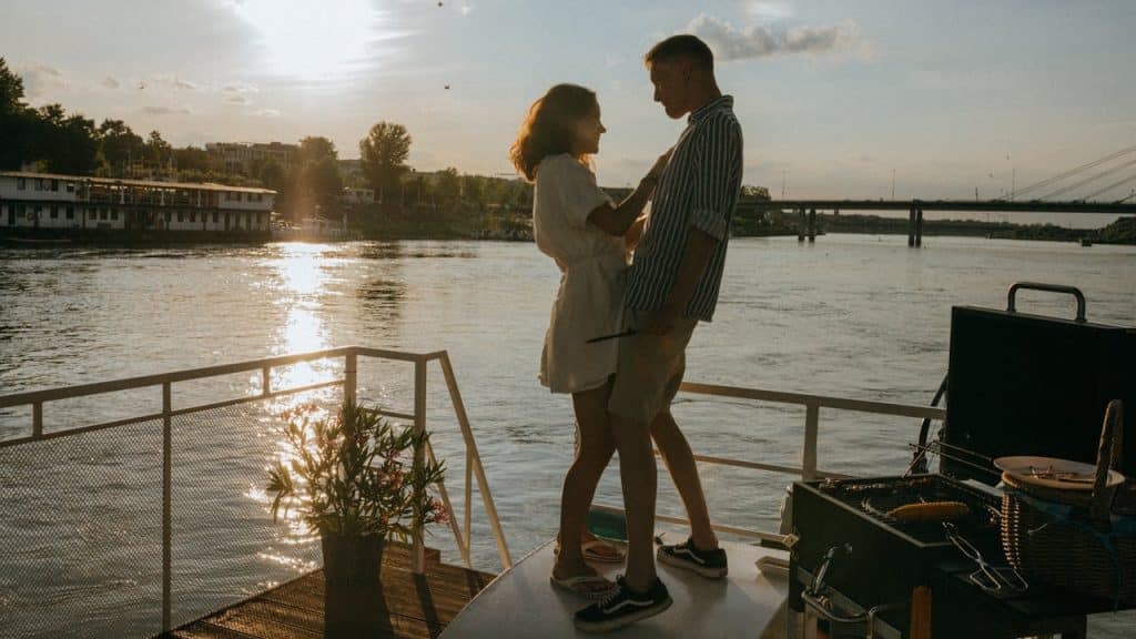 Romantic couple embracing on a boat deck at sunset with a river and cityscape in the background.