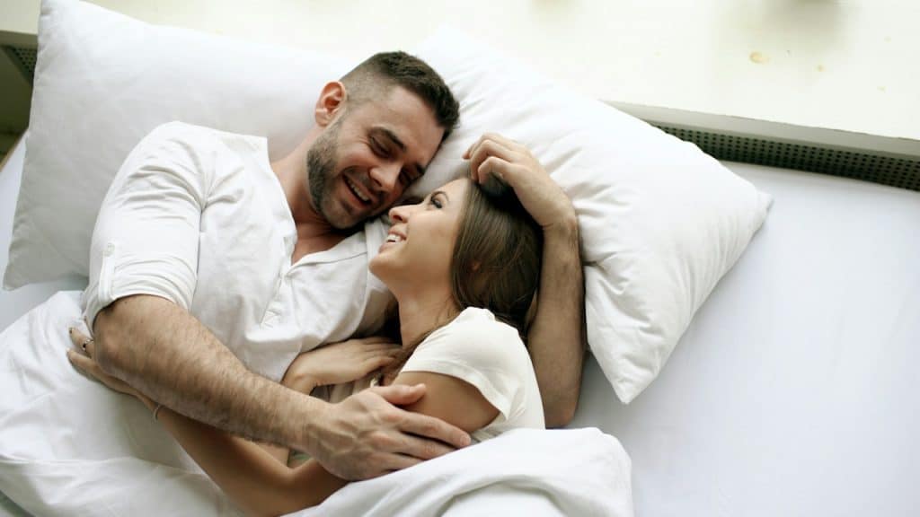 A couple embracing in bed are smiling at each other.
