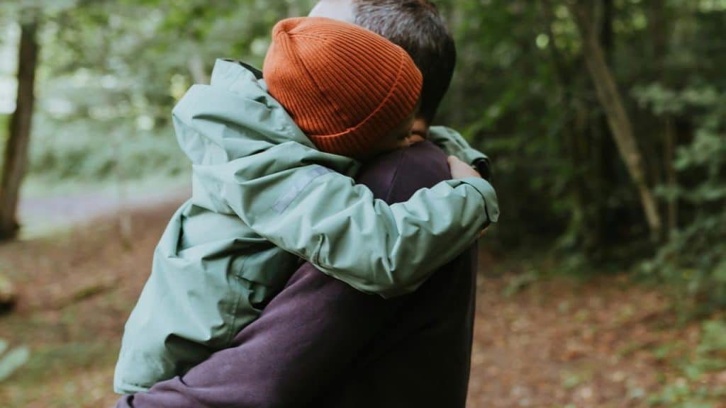 A person wearing a beanie and jacket hugs another person in a forest.