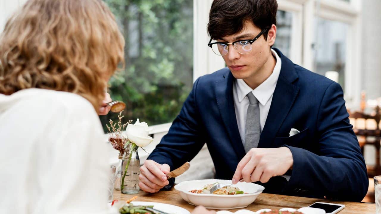 A man in a suit and glasses eating a meal across a table from a woman.