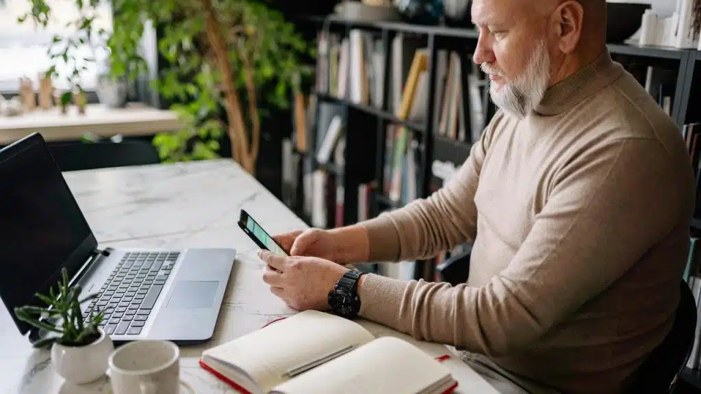 A man sitting at a desk using his phone beside an open laptop and notebook.
