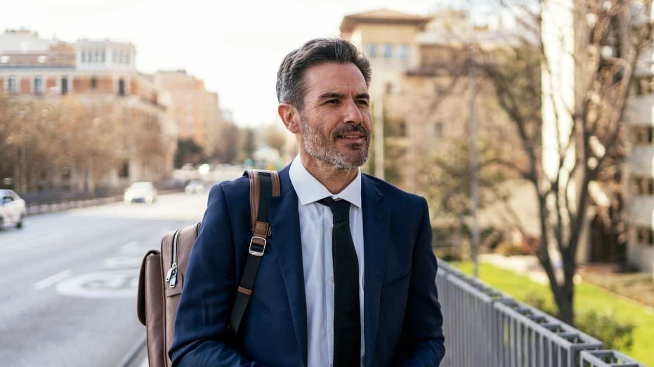 A man in a suit with a backpack is walking outdoors in an urban area.