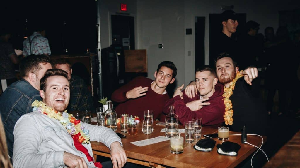 A group of young men, some wearing leis, are sitting around a table in a dimly lit setting, smiling and posing for the camera.