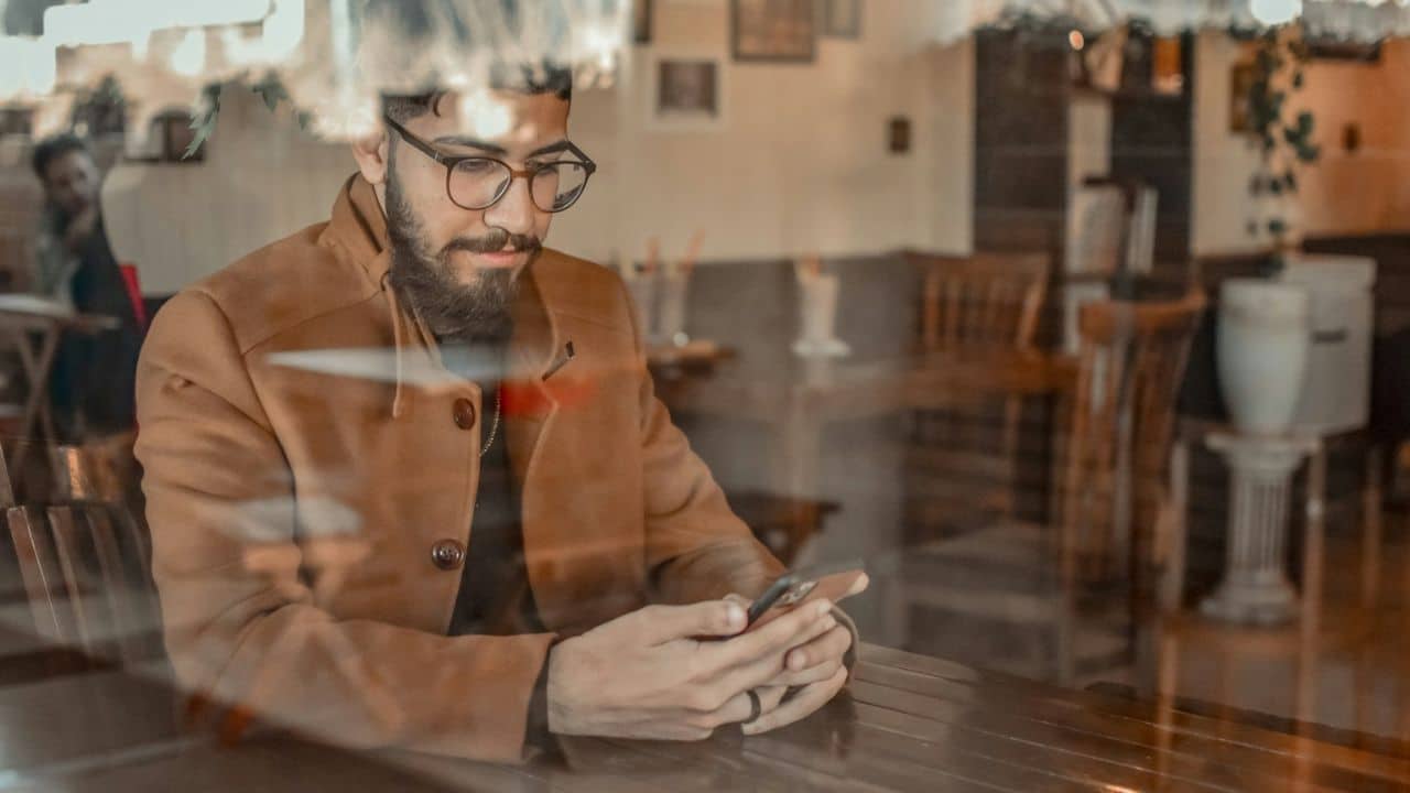 A bearded man in glasses and a brown coat looks at his phone in a cafe window reflection.