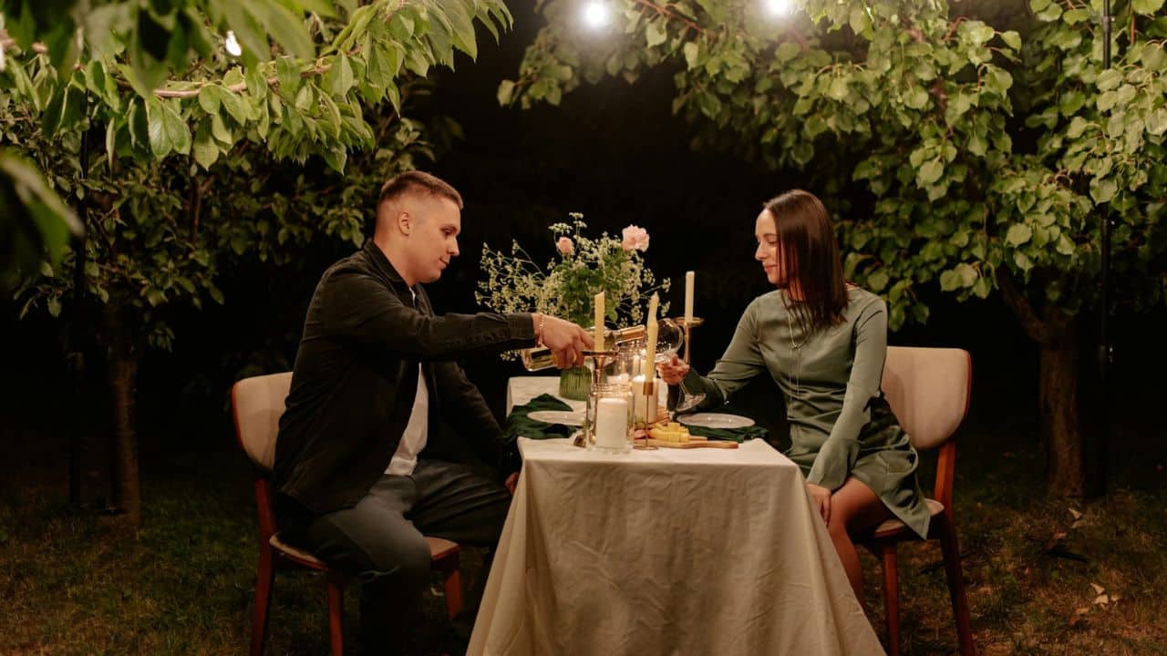 A man is pouring wine for a woman during a romantic, candlelit dinner outdoors at night.