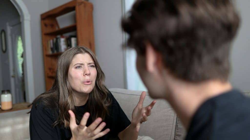 Woman sitting on a couch, passionately arguing and gesturing with her hands towards a blurred man.