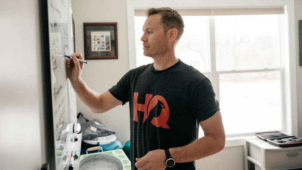 A man in a dark t-shirt writes on a white board next to a window.