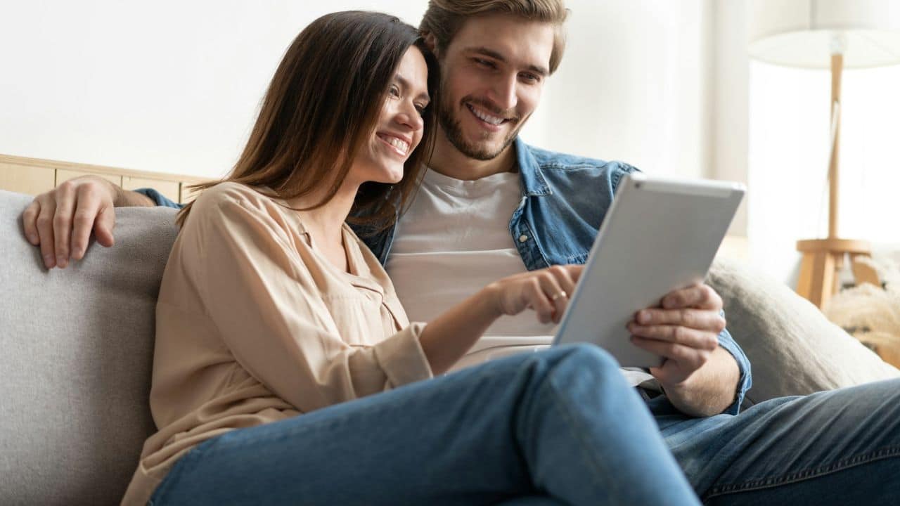 A smiling couple sits on a couch looking at and pointing at a tablet.