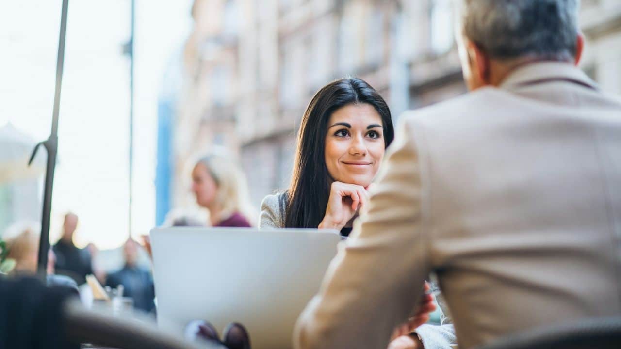 A woman smiling sits across from a man outdoors, with a laptop between them.