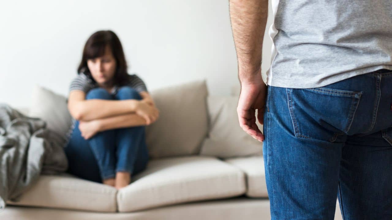 A man stands facing away as a sad woman sits curled up on a couch behind him.