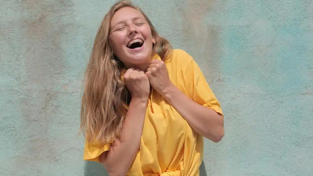 A joyful woman in a yellow shirt laughing with her hands clasped near her face.