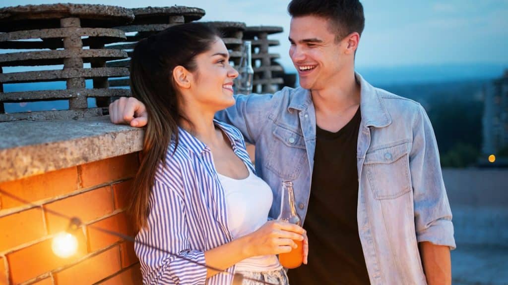 A smiling couple is standing close together on a rooftop patio at dusk, looking at each other.