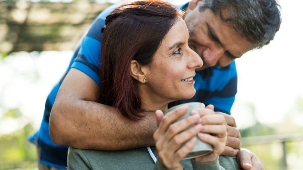 A man wrapping arms around a woman’s shoulder who is having coffee.