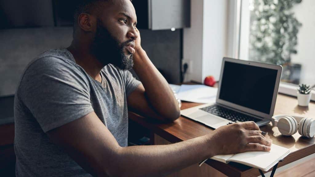 A man sitting at a desk with a laptop, resting his head on his hand and looking tired.