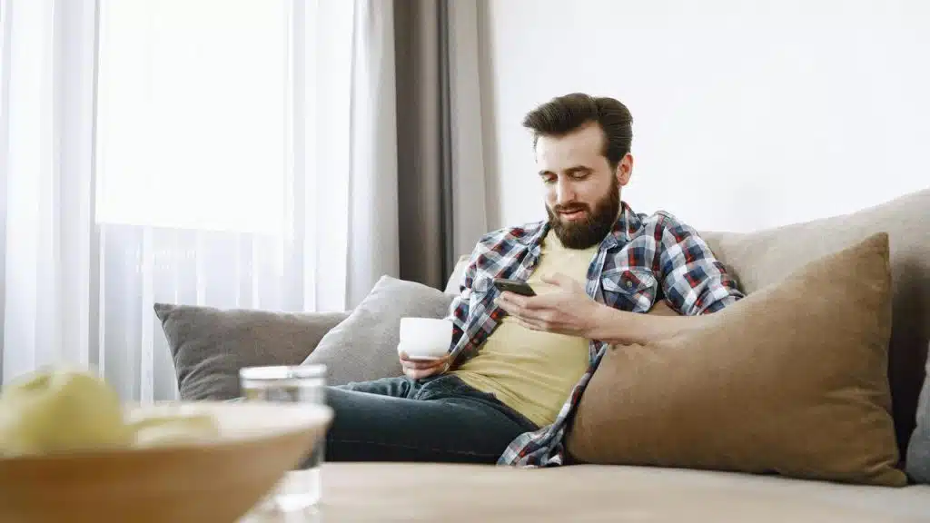 A man sitting on a couch holding a cup and looking at his phone.