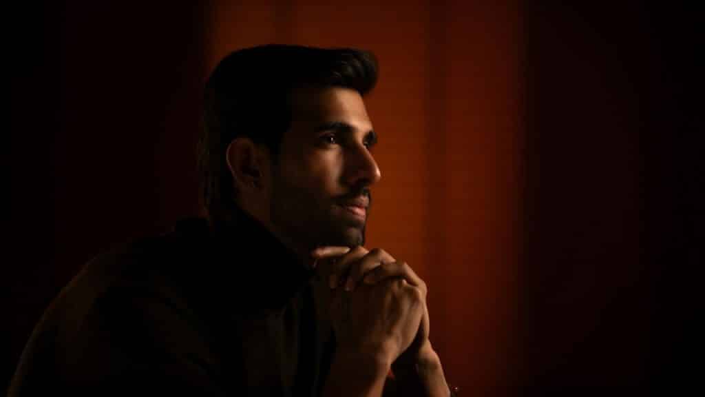 A man in a turtleneck, chin resting on clasped hands, looks to the right in dim light.