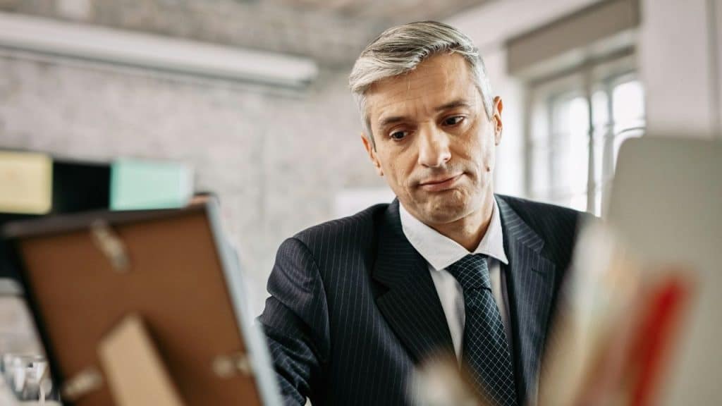 Middle-aged man in a suit and tie looks down sadly at a framed object on his desk.