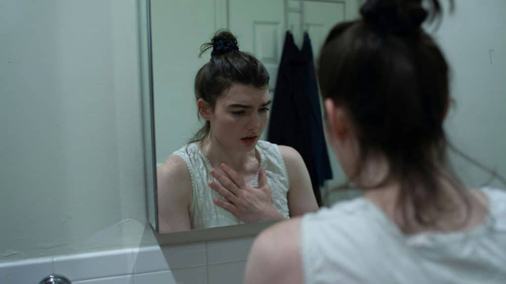 Woman with hair in a bun looking distressed at her reflection in a mirror.