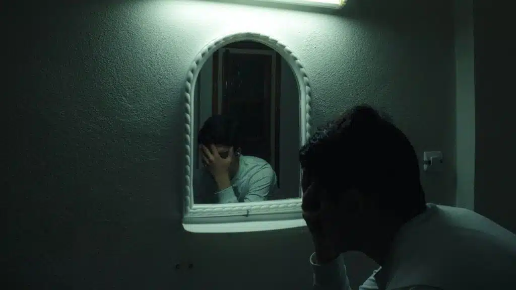 A man in a dark room holds his head, reflected in a small, illuminated mirror.