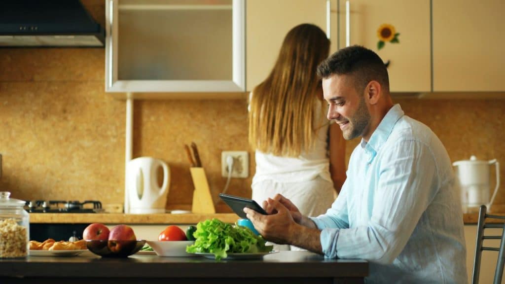 A man smiles while using a tablet at a table; a woman is in the background kitchen.